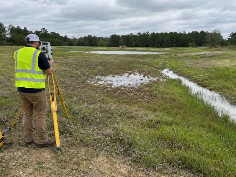 Topographic Surveys Warner Robins Georgia | Warner Robins Land Surveying Surveyor in a high-visibility vest uses a theodolite/level on a tripod in a wet, grassy field with a small water channel nearby.